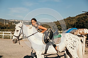 Beautiful woman with dark hair in elegant sportive suit posing near white horse in stable
