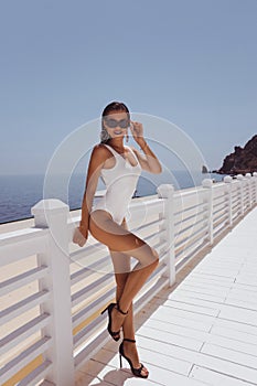 beautiful woman with dark hair in elegant white swimming suit and sunglasses posing on the terrace with cup of coffee