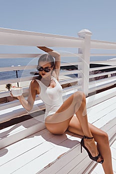 beautiful woman with dark hair in elegant white swimming suit and sunglasses posing on the terrace with cup of coffee