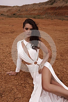 beautiful woman with dark hair in luxurious white dress with accessories posing in desert in Cyprus