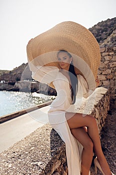 Beautiful woman with dark hair in luxurious white dress and hat posing in beautiful landscape with sea view