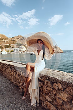 Beautiful woman with dark hair in luxurious white dress and hat posing in beautiful landscape with sea view