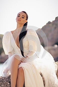 Beautiful woman with dark hair in luxurious white dress posing in beautiful landscape with sea view