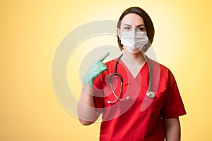 Beautiful woman doctor with stethoscope, wearing red scrubs wears a protective mask, with her finger pointed