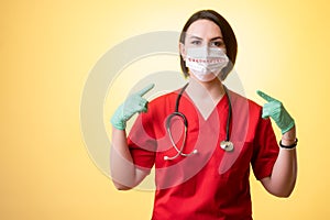 Beautiful woman doctor with stethoscope, wearing red scrubs wears a protective mask, with her fingers pointed