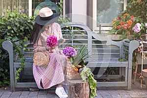 Beautiful woman in a dress with purchases, sits on a bench in the city street. A large basket of vegetables and flowers in the