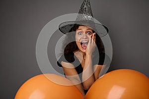 Beautiful woman dressed in black and wizard hat, looks at camera, expresses stupefaction and surprise. Halloween concept on gray photo