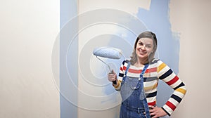 Beautiful woman dressed in overalls, smiling at camera with blue paint roller in her hand against half-painted white wall