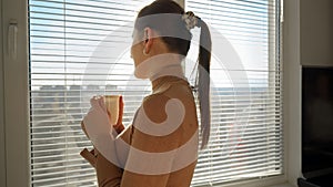 Beautiful woman drinking coffee and opening window blinds. People in morning, beautiful cityscape, having break at office