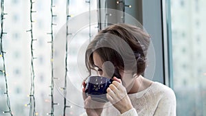 Beautiful woman drinking tea and looking out window with garlands. Winter warm comfort. Woman looks out window waiting