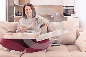 Beautiful woman eating pizza and watching TV holding remote control at home, orange tone