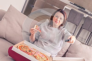 Beautiful woman eating pizza and watching TV holding remote control at home, orange tone