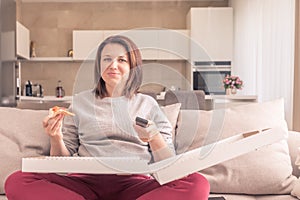 Beautiful woman eating pizza and watching TV holding remote control at home, orange tone