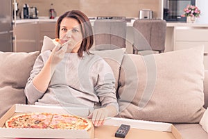 Surprised woman eating pizza and watching TV with remote control at home, warm tone