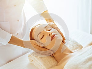 Beautiful woman enjoying facial massage with closed eyes in sunny spa center. Relaxing treatment concept in medicine