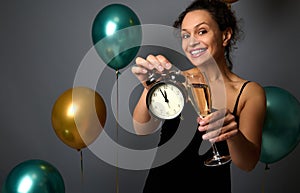 Beautiful woman in evening black dress holds a flute of sparkling wine and shows an alarm clock isolated over gray background with