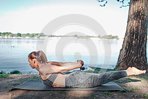 Beautiful woman is excercising at the river shore. She is keeping hands together above back and balancing with her legs