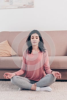Beautiful woman with eyes closed in Lotus Pose practicing meditation in Living Room at home.