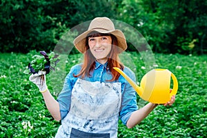 Beautiful woman farmer holds a bunch of basil and watering garden in a straw hat and surrounded by the many plants in her