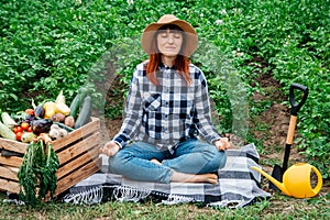 Beautiful woman farmer in a straw hat meditating while sitting on a blanket near fresh organic vegetables in a wooden box against
