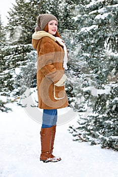 Beautiful woman full length on winter outdoor, snowy fir trees in forest, long red hair, wearing a sheepskin coat