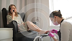 Beautiful woman getting professional pedicure in spa beauty salon. Master is cleaning her nails. Client is chatting on