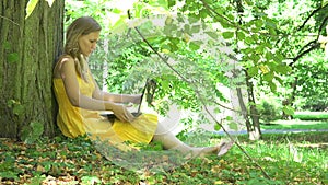 Beautiful woman girl in dress sit near tree trunk and start using laptop computer in park. 4K