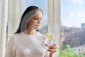 Beautiful woman with glass of water with lemon at home near window