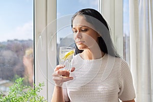 Beautiful woman with glass of water with lemon at home near window