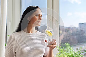 Beautiful woman with glass of water with lemon at home near window