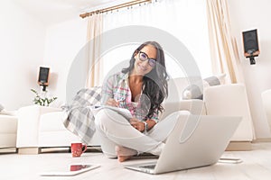 Beautiful woman in glasses smiling with a tilted head while sitting on the living room floor working on a pc and notepad