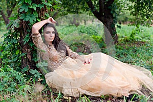 Beautiful woman in gold dress is sitting leaning on a tree