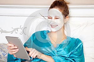 Beautiful woman with green nightie and white facial mask resting on bed and holding tablet before sleeping.