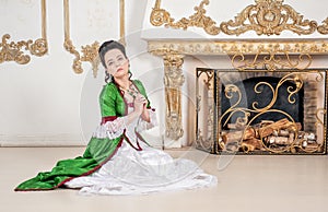 Beautiful woman in green rococo style medieval dress sitting on the floor near fireplace and praying