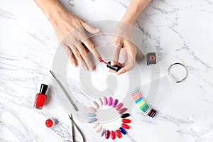 Beautiful woman hands painting nails with red nail polish on marble table with manicure set on it.
