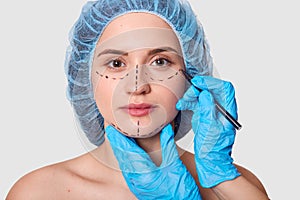 Beautiful woman has marked arrows under eyes. Lady wants to improve her appearance. Plastic surgeon with blue gloves draws black