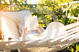 Beautiful woman with hat reading book while relaxing in hammock in the garden