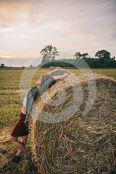 Beautiful woman in hat relaxing on haystack in summer evening field. Tranquility in countryside
