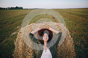 Beautiful woman in hat relaxing on haystack in summer evening field. Tranquility in countryside