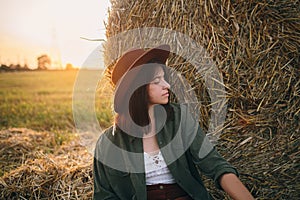 Beautiful woman in hat relaxing at haystacks enjoying evening sunset in summer field. Tranquility