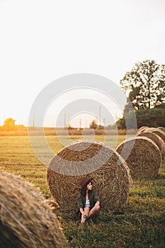 Beautiful woman in hat relaxing at haystacks enjoying evening sunset in summer field. Tranquility