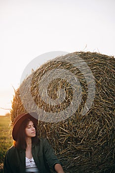 Beautiful woman in hat relaxing at haystacks enjoying evening sunset in summer field. Tranquility