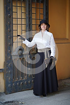 a beautiful woman in a hat and vintage clothes stands at the gate
