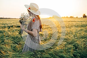 Beautiful woman in hat with wildflowers enjoying sunset in barley field. Atmospheric tranquil moment, rustic slow life. Stylish