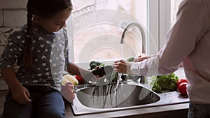 Beautiful woman with her four year old daughter is washing vegetables in the kitchen sink.