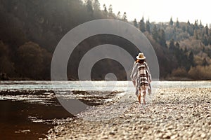 Beautiful woman hipster walking on river beach in mountains, wearing hat and poncho, boho travel concept, space for text