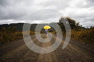 Beautiful woman hold yellow umbrella and walks on a country road under rain