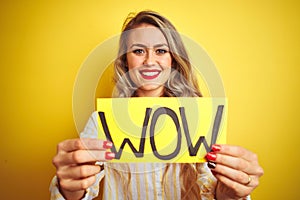 Beautiful woman holding amazed wow surprise banner over isolated yellow background with a happy face standing and smiling with a