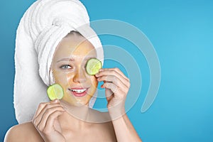 Beautiful woman holding cucumber slices near her face with natural mask against color background
