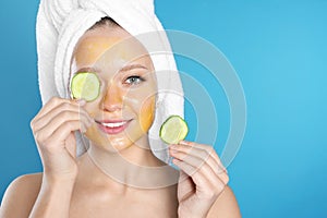 Beautiful woman holding cucumber slices near her face with natural mask against color background